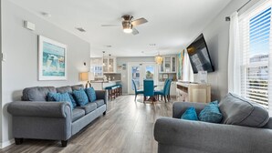 Una sala moderna con dos sofás grises, un comedor con sillas azules y una cocina con un televisor montado en la pared.