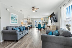 A modern living room with two grey sofas, a dining area with blue chairs, and a kitchen with a TV mounted on the wall. - New Kulula WEST-  3 BR, Sleeps 9, Heated Salt Water Pool, Bikes & moments from the beach (Santa Rosa Beach)
