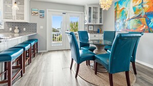 Una cocina moderna con una isla de vidrio, taburetes de bar azules y una zona de comedor con sillas verde agua y una mesa de vidrio.