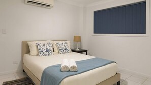 A hotel room with a bed, a nightstand, a lamp, and a window with curtains. - Pebbly Beach Estate | Sea View (Forster)