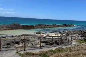 Una zona costera rocosa con aguas azul turquesa, una playa de arena y una barandilla metálica en primer plano.