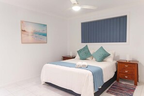 Un dormitorio con cama, dos mesitas de noche, ventilador de techo, ventana con persianas, un cuadro en la pared y alfombra en el suelo.