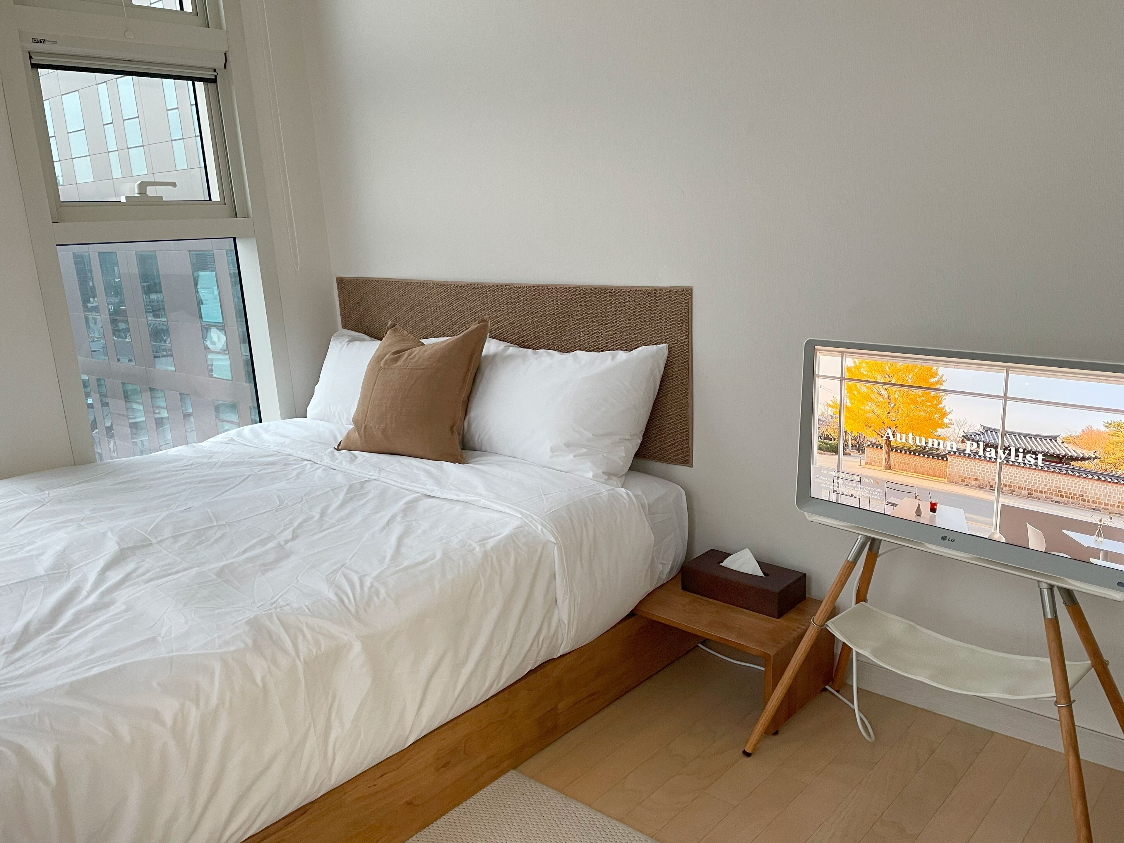 A neatly made bed with white linens and two pillows, a wooden headboard, a bedside table with a tissue box, and a view of a traditional building through a window.