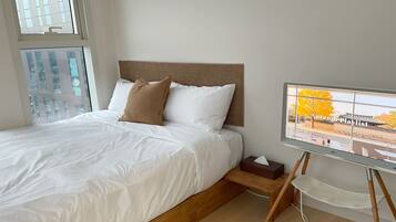 A neatly made bed with white linens and two pillows, a wooden headboard, a bedside table with a tissue box, and a view of a traditional building through a window.