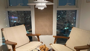 Una habitación con dos sillas de mimbre, una mesa con comida y bebidas, y vistas a la ciudad a través de amplias ventanas.