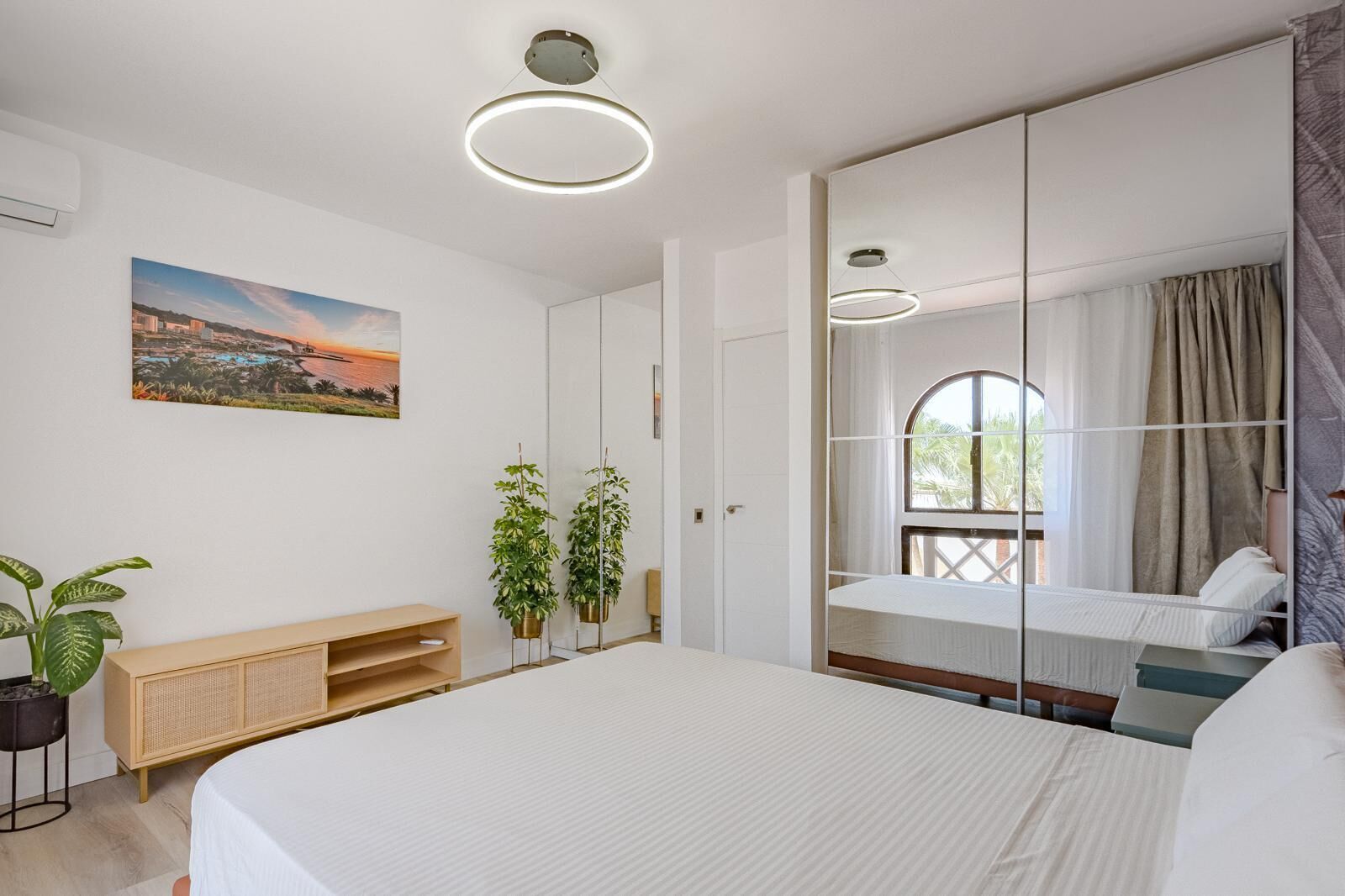 A bedroom with a bed, bedside table, potted plants, a painting, and a window with curtains.