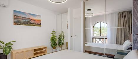 A bedroom with a bed, bedside table, potted plants, a painting, and a window with curtains.