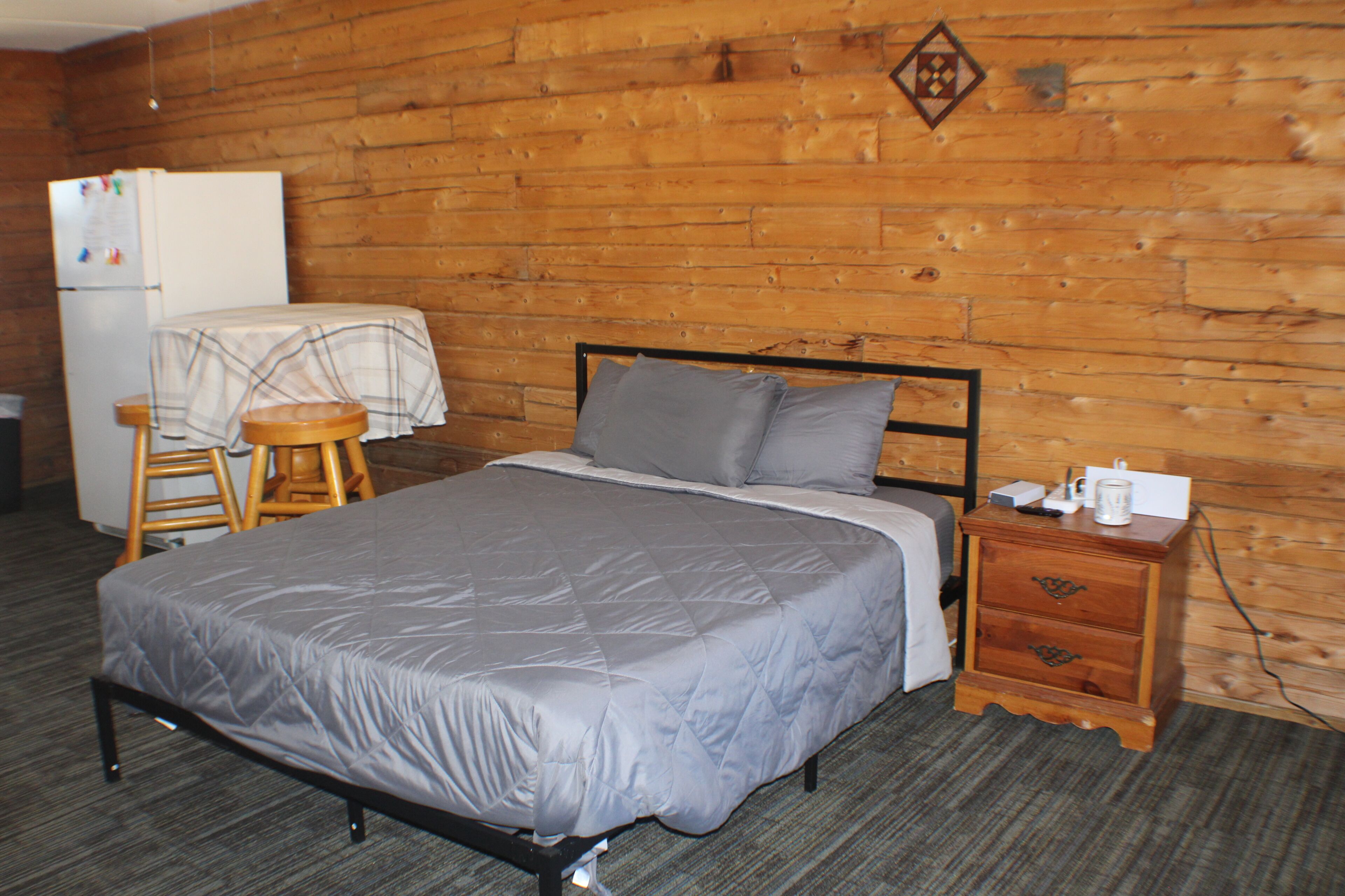 A room with a bed, wooden walls, a small wooden nightstand, a chair, and a refrigerator.