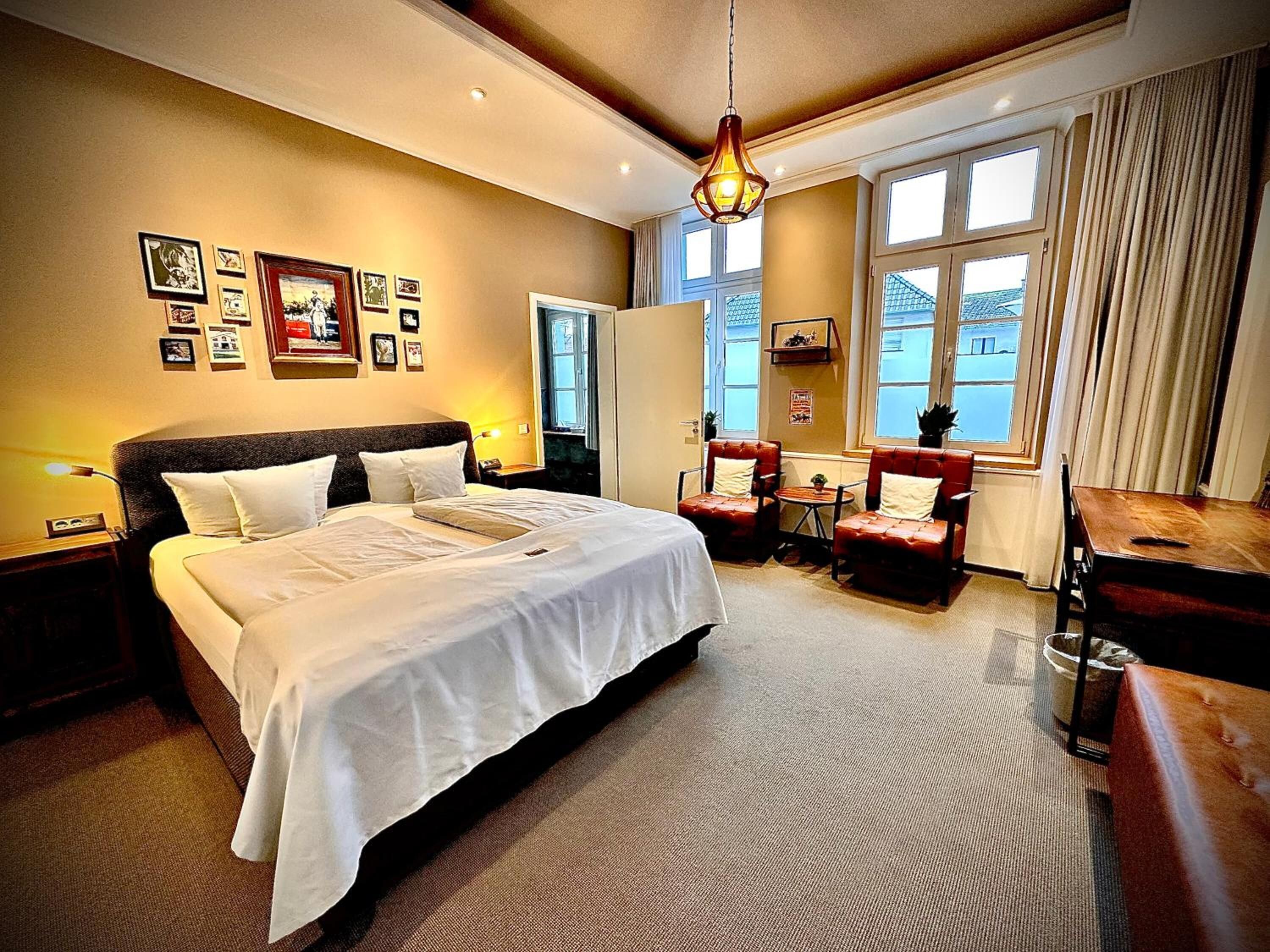 A hotel room with a large bed, a desk, a chair, a small table, and a window with curtains.