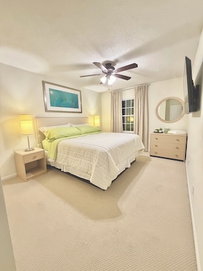 Une chambre à coucher comprenant un lit, des tables de chevet, un ventilateur de plafond, une commode, un miroir et une fenêtre avec des rideaux.