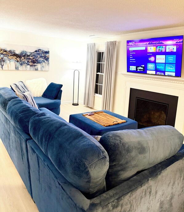 Un salon moderne avec un grand canapé bleu, une table basse en bois et un téléviseur à écran plat fixé au-dessus d’une cheminée.