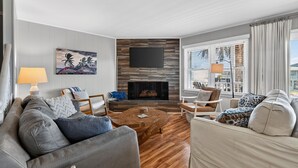 A modern living room with a fireplace, comfortable seating, and a large window. - Sunsets, Sand & Boat-Ready Getaway (Holden Beach)