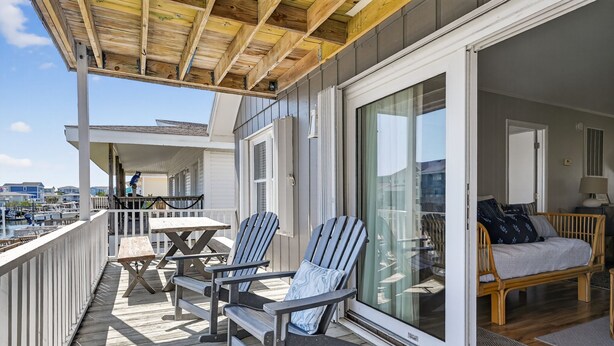 A wooden deck with outdoor furniture, a sliding glass door leading to a room with a sofa and a lamp. - Sunsets, Sand & Boat-Ready Getaway (Holden Beach)