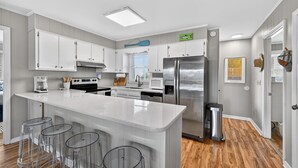 A modern kitchen with white cabinets, a large island with bar stools, stainless steel appliances, and a wooden floor. - Sunsets, Sand & Boat-Ready Getaway (Holden Beach)