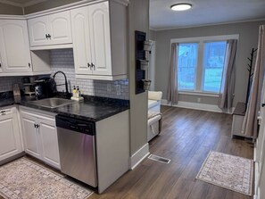 A modern kitchen with white cabinets, a black countertop, and a dishwasher. There is a window with blinds and a rug on the floor. - Serene 1-bedroom house in welcoming Slippery Rock (Slippery Rock)