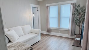 A modern living room with a beige sofa, a small side table, a potted plant, and a window with curtains. - Serene 1-bedroom house in welcoming Slippery Rock (Slippery Rock)