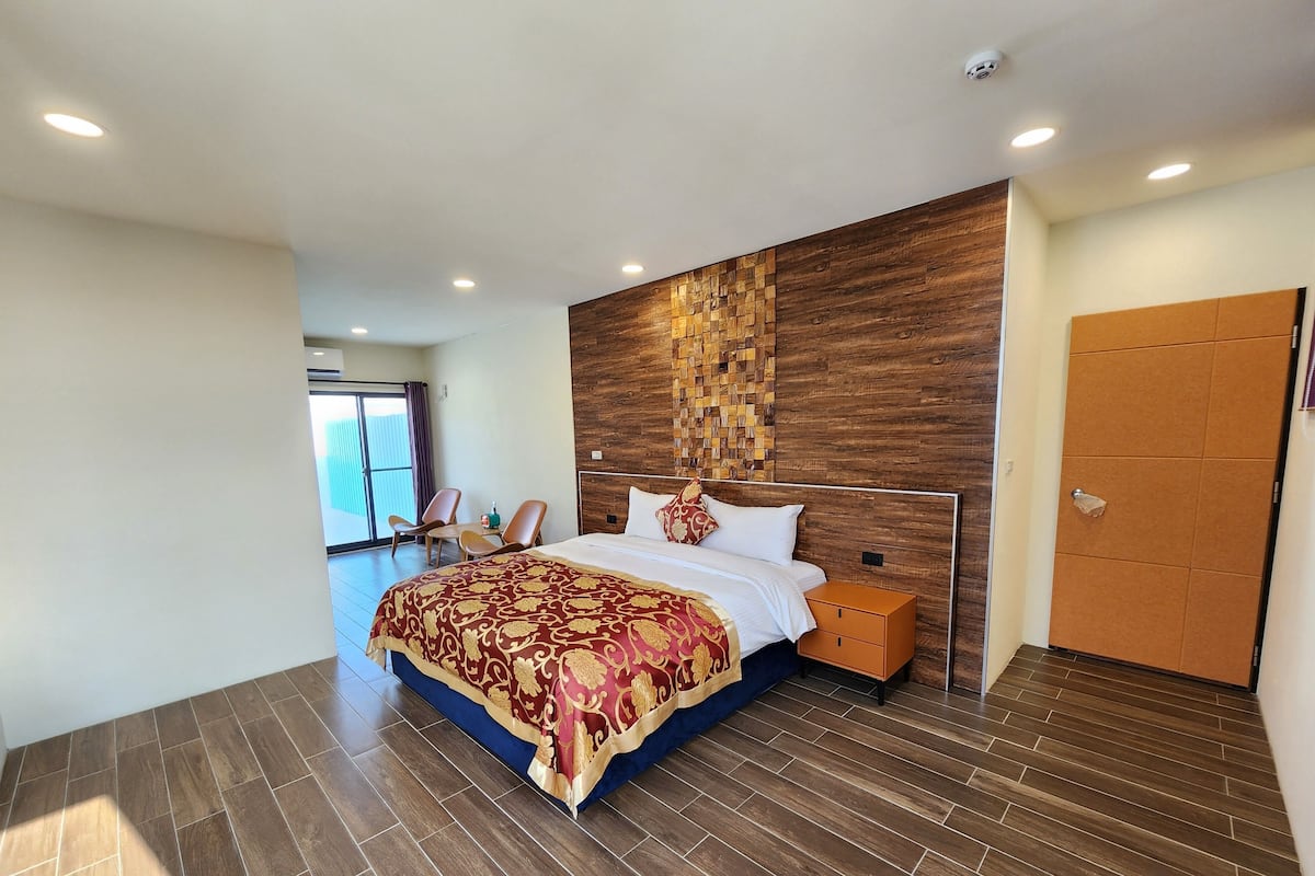 A modern hotel room with a large bed, a wooden accent wall, a sliding door, and a view of the outdoors.