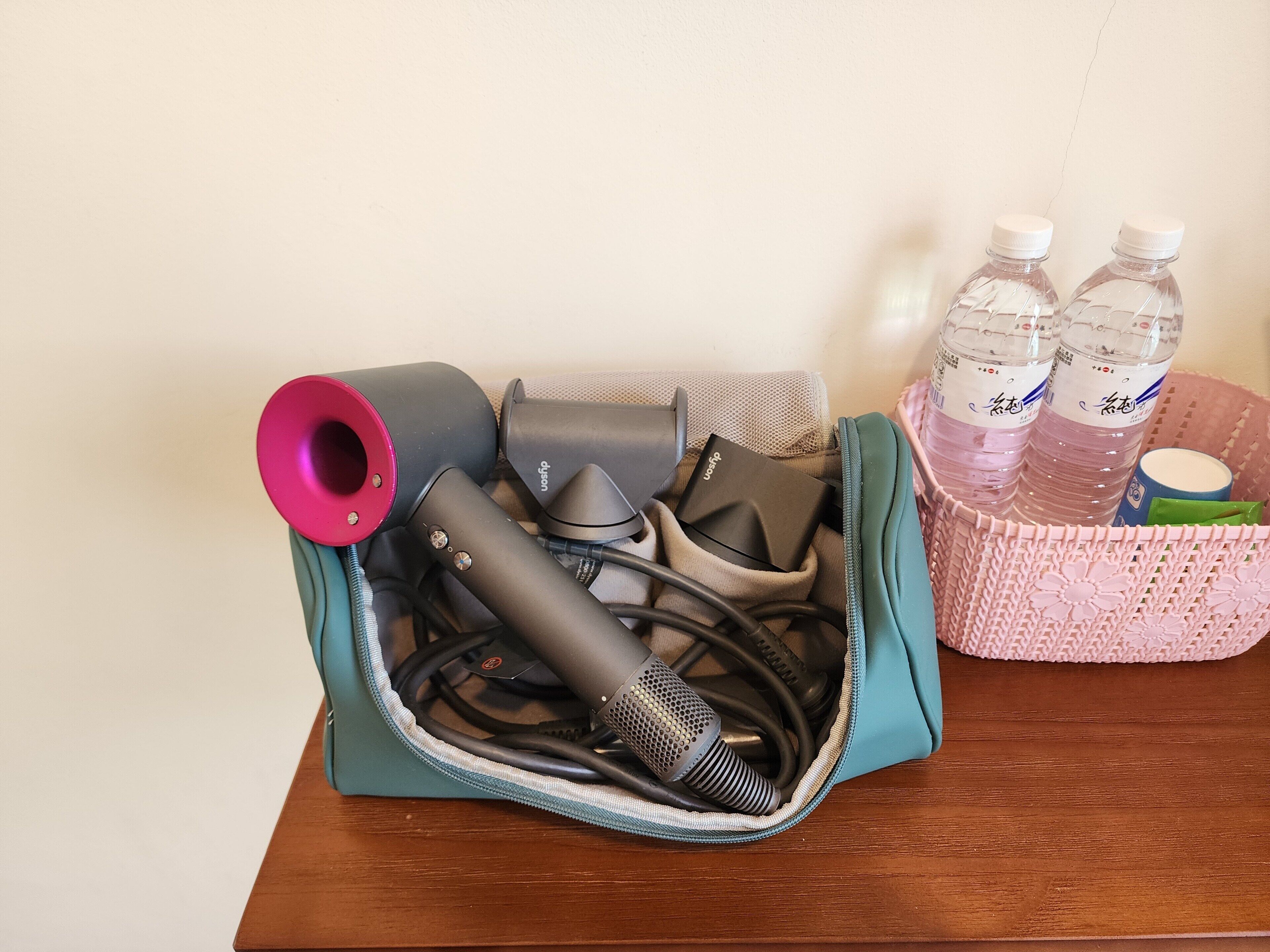 A teal bag with a microphone and hair styling tools on a wooden surface, next to a pink basket with water bottles.