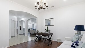 Una sala moderna con piso de madera oscura, paredes blancas, un sofá blanco, una mesa de comedor con sillas, una araña y una cocina al fondo.
