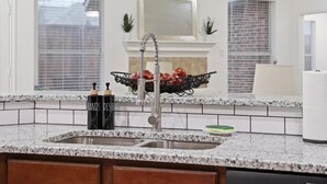 Cocina con encimeras de granito, un lavamanos de acero inoxidable, un dispensador de jabón negro y una cesta de tomates.