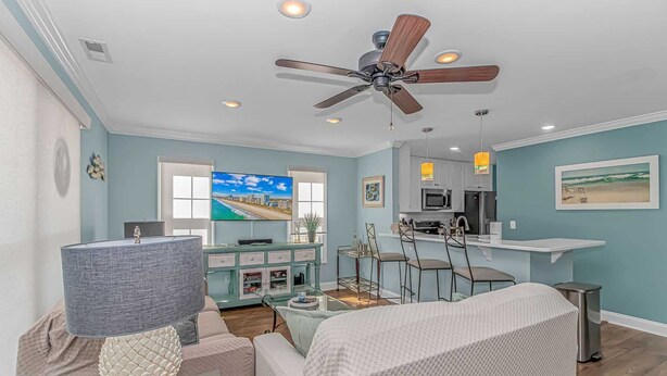 A modern living room with a ceiling fan, a TV, a kitchen area, and a dining space. - Charleston Village 201 (North Myrtle Beach)