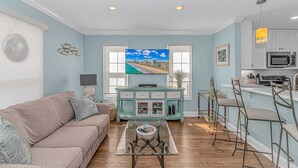 A living room with a sofa, a coffee table, a TV, and a kitchen area with a bar and a microwave. - Charleston Village 201 (North Myrtle Beach)