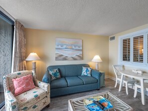 A living room with a sofa, armchair, and table, a painting on the wall, and a window with shutters. - Beach Colony 603 (Myrtle Beach)