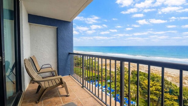 A balcony with a view of the ocean, a chair, and a glass door. - Beach Colony 603 (Myrtle Beach)