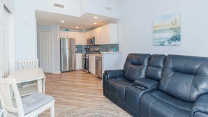 A modern living room with a leather sofa, a dining table with chairs, and a kitchen area with white cabinets and stainless steel appliances. - Ocean Escape 1227 (Myrtle Beach)