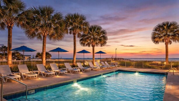 A swimming pool with lounge chairs and umbrellas, surrounded by palm trees. - Ocean Escape 1227 (Myrtle Beach)