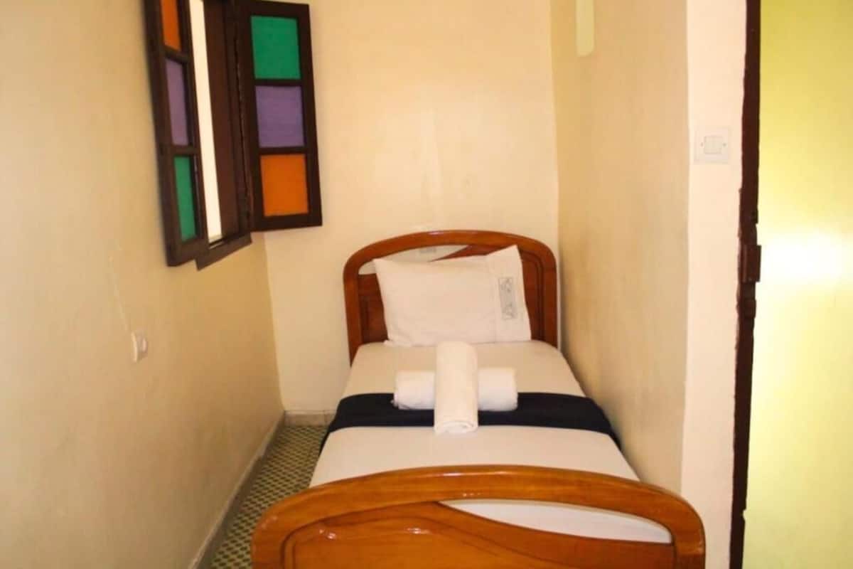 A narrow, single-bed room with a wooden headboard, a white pillow, and a rolled towel on the bed.