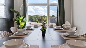 Ruokapöytä on katettu valkoisilla lautasilla ja kulhoilla, siinä on myös ruukkukasvi, ja ikkunasta avautuu näkymä metsään.
