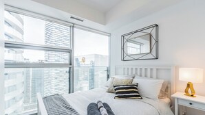 Quarto de hotel com uma janela grande, uma cama com almofadas, uma mesa de cabeceira com abajur e um espelho geométrico na parede.