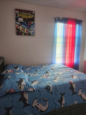 Une chambre à coucher avec un lit recouvert d’une couverture bleue ornée de motifs de requins, une affiche de Spider-Man accrochée au mur et une fenêtre avec des rideaux rouges et bleus.