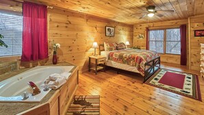A wooden cabin bedroom with a large bed, a wooden floor, a bathroom with a bathtub, and a window with curtains. - FALL FOLIAGE RETREAT! 1BR+Loft Cabin w/Hot Tub & Arcade (Sevierville)