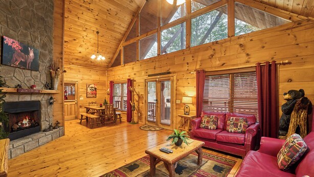 A spacious log cabin interior with a fireplace, red leather sofa, wooden dining table, and large windows. - FALL FOLIAGE RETREAT! 1BR+Loft Cabin w/Hot Tub & Arcade (Sevierville)