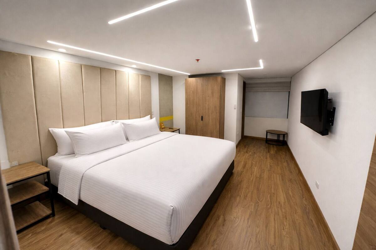 A modern hotel room with a large bed, a wooden nightstand, a small table, and a flat-screen TV mounted on the wall.