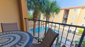 A balcony with a round table and chairs, a pool, and a building with balconies in the background. - Spacious 3-bedroom condo in the heart of Tybee Island. Easy beach access. (Tybee Island)