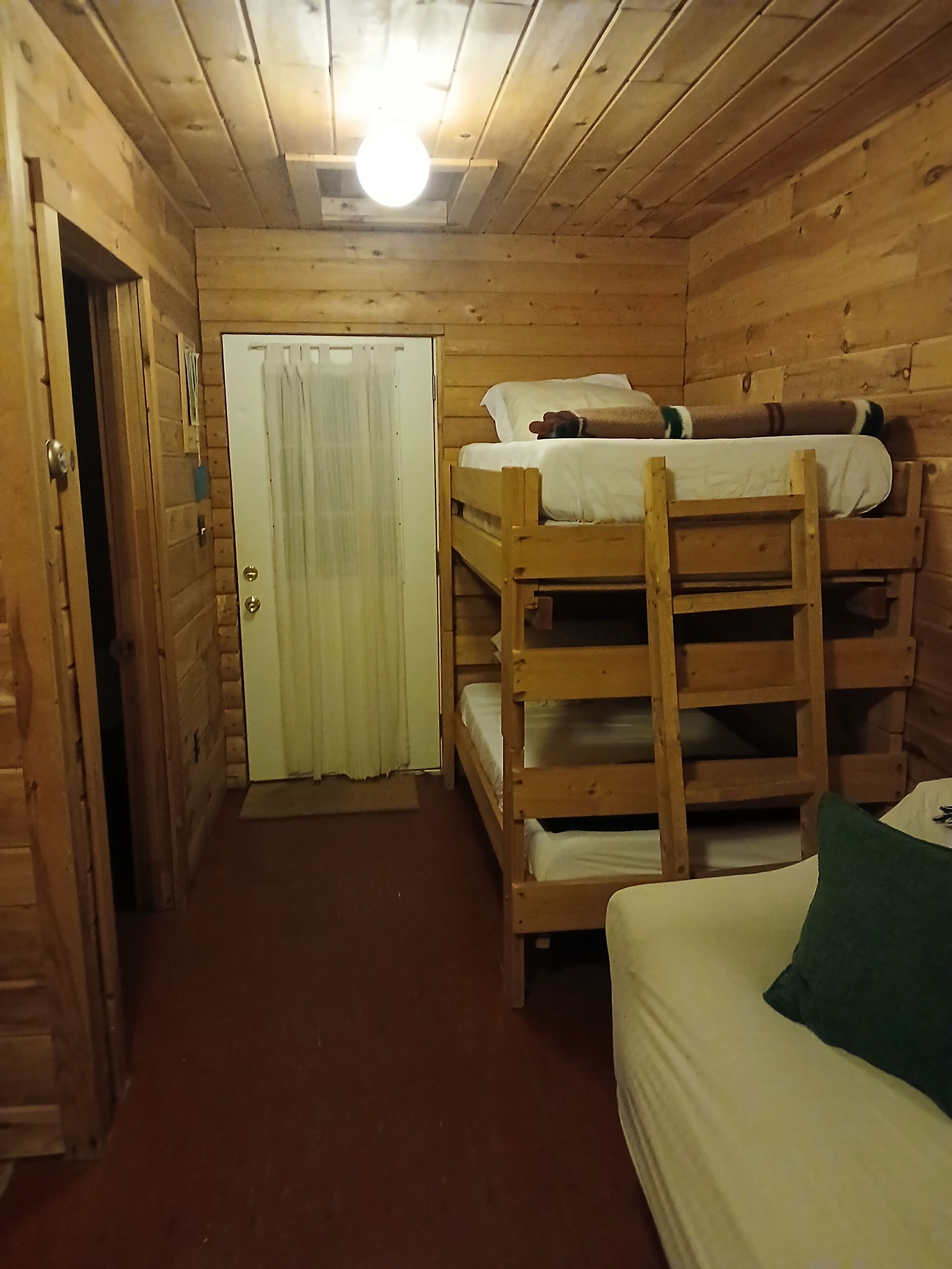 A wooden cabin room with bunk beds, a door with a curtain, and a light fixture on the ceiling.