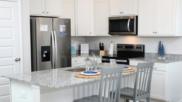 Cocina moderna con armarios blancos, electrodomésticos de acero inoxidable, encimera de granito y una isla central con dos sillas.