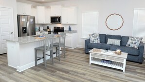 Una cocina moderna con armarios blancos, un refrigerador de acero inoxidable y un microondas. Hay un sofá gris con cojines estampados y una pequeña mesa de centro blanca con objetos decorativos.