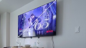 Moderni olohuone, jonka seinälle on asennettu litteänäyttöinen televisio, jolta näkyy Netflix-sarja Bridgerton. Huoneessa on kaksi valkoista pöytää tuoleineen, pieni matto ja puulattia.