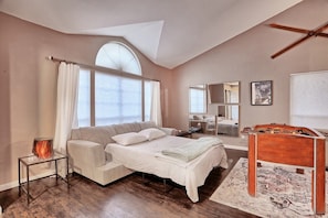 A bedroom with a bed, a bedside table, a lamp, a window with curtains, a ceiling fan, a mirror, and a wooden dresser.