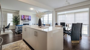 Een moderne keuken met een centraal eiland, een eethoek met een donkere stoel en een groot raam met jaloezieën.