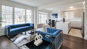 Een moderne woonkamer met een blauwe bank, een witte fauteuil en een eethoek, met een keuken op de achtergrond.