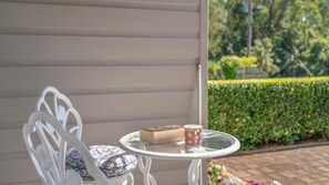 A white metal chair with a cushion, a small round table with a book and a patterned mug, and a hedge in the background. - Shanti for 10 at Berry (Bellawongarah)
