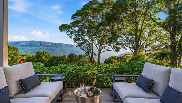 Une terrasse avec deux canapés gris, une table en métal avec une bouteille et des verres, et une vue sur un paysage montagneux.