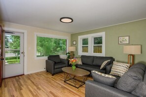 Ein Wohnzimmer mit zwei Sofas, einem Couchtisch und einer Lampe. Fenster bieten Blick ins Grüne und eine Tür befindet sich links.