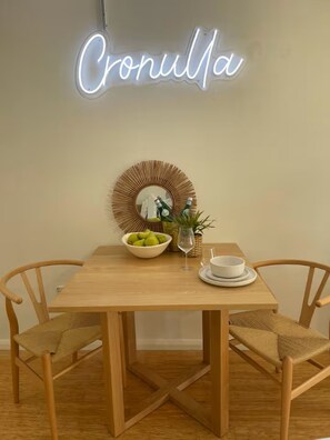 A dining table with a neon sign reading "Cronulla," a bowl of green apples, a glass, and a white bowl on a white plate. - A Cozy 1BR in Cronulla – Walk to Beach & Shops U4 (Cronulla)