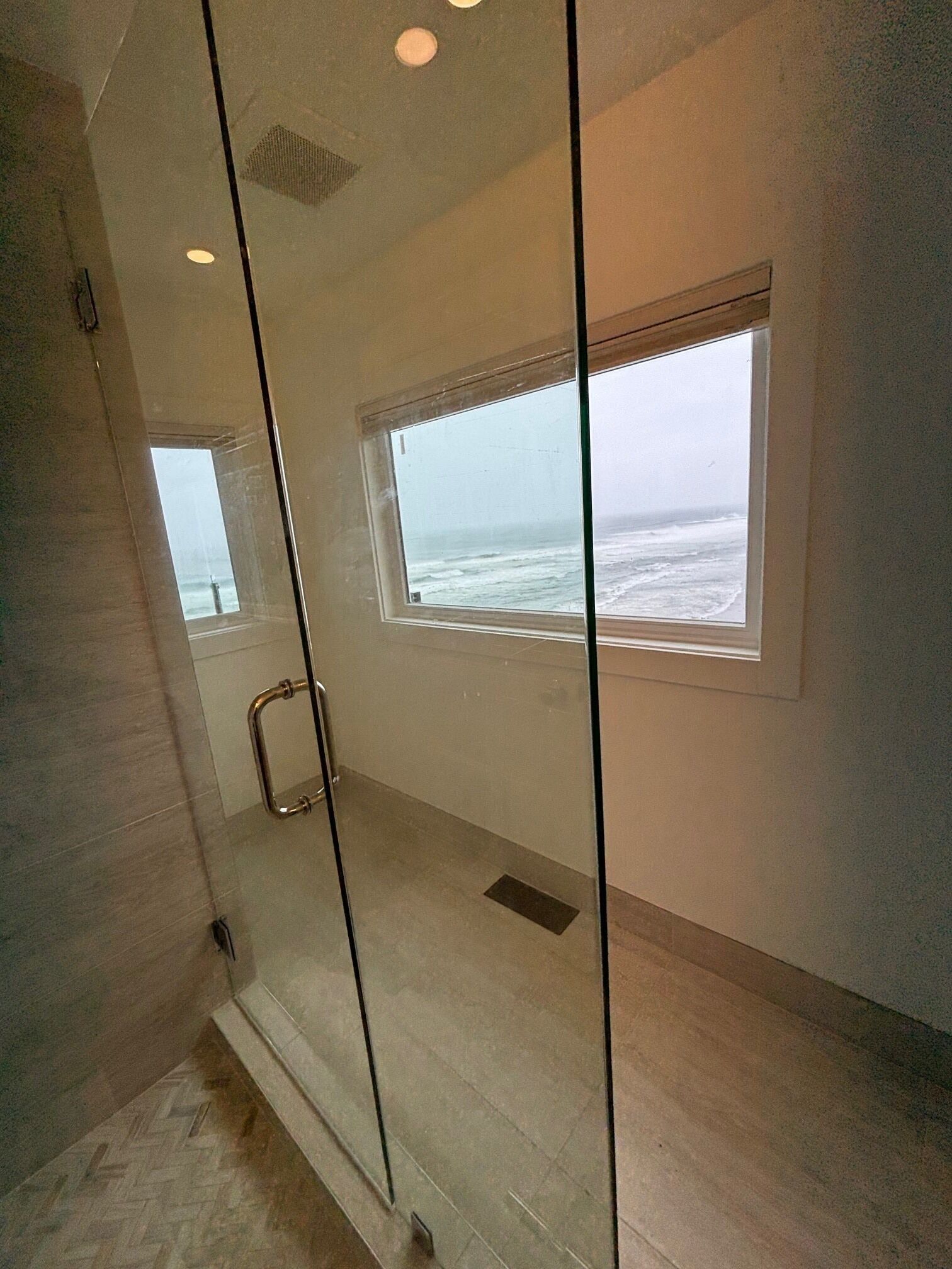 Une salle de bain moderne dotée d’une douche à parois de verre, d’une petite fenêtre donnant sur l’océan et d’un sol décoré d’un motif en chevrons.
